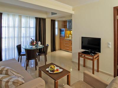 Hotelový obývací pokoj s pohovkou, stolkem s ovocem, televizí a jídelním stolem u okna.