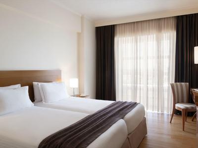 Světý pokoj hotelu s dvěma jednolůžky, tmavými závěsy a dřevěným křeslem u okna.