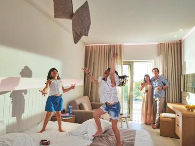 Děti a rodiče radostně skáčou na posteli v hotelovém pokoji.