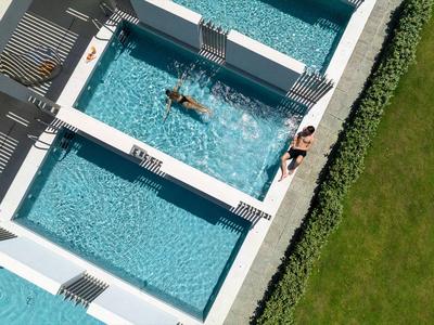 Drei Personen entspannen in einem Hotelpool mit klar blauem Wasser neben einem gepflegten Rasen.