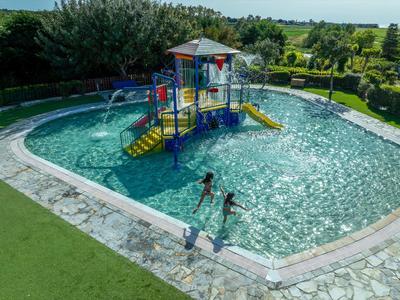 Mělý bazén s vodním hřištěm a skluzavkou, obklopený zeleným trávníkem.