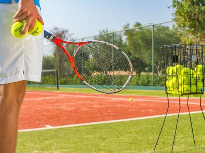 Person hält Tennisbälle und Schläger auf einem Tennisplatz mit Ballkorb im Hintergrund.