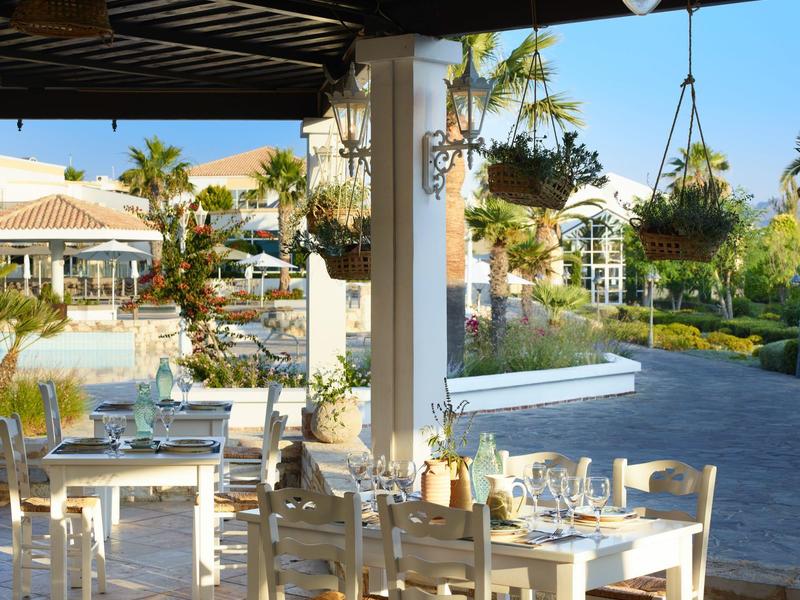 Restauranční terasa s bílými stoly a židlemi pod přístřeškem, zdobená závěsnými rostlinami.