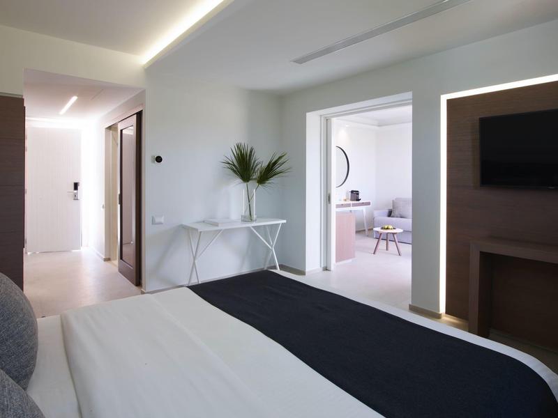 Moderní pokoj s manželskou postelí, stolem s rostlinou a výhledem na světlý obývací pokoj.