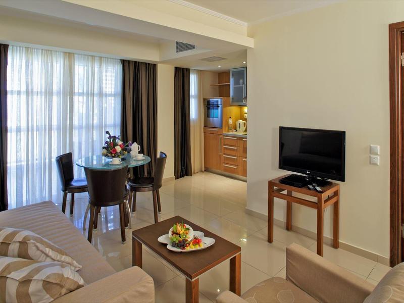 Hotelový obývací pokoj s pohovkou, stolkem s ovocem, televizí a jídelním stolem u okna.