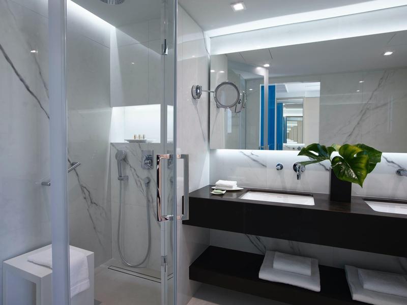 Moderní hotelová koupelna se skleněným sprchovým koutem a osvětleným zrcadlem.