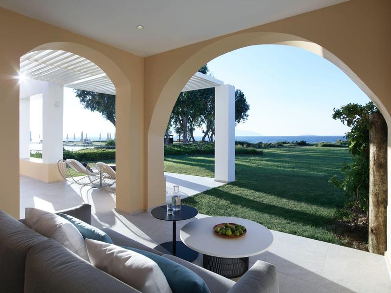 Hotelová terasa s výhledem na zahradu a moře, béžové oblouky a moderní bílé posezení.