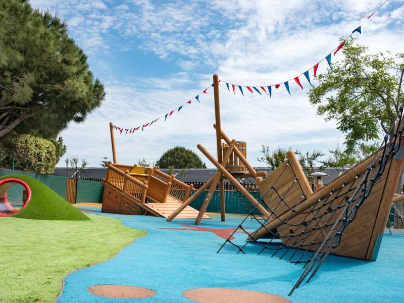 Moderní venkovní dětské hřiště s dřevěnými konstrukcemi a barevným povrchem pod modrou oblohou.