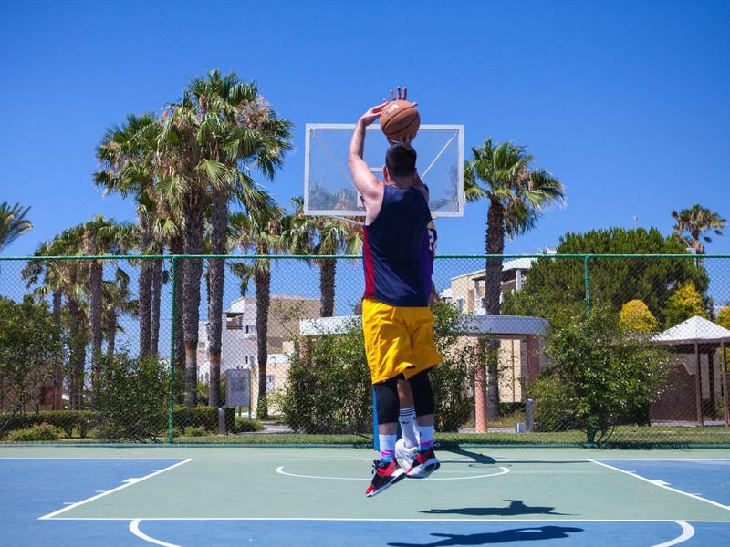 Muž hrající basketbal venku na hřišti obklopeném palmami pod modrou oblohou.