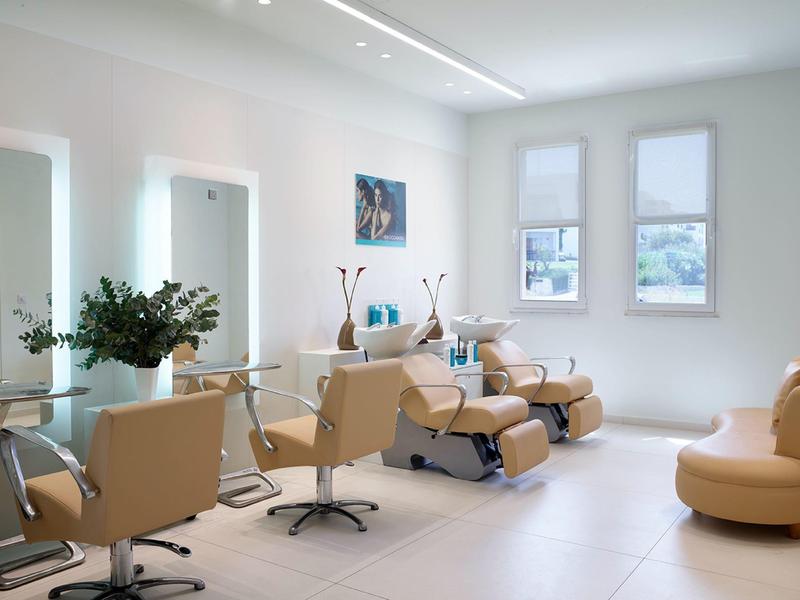 Moderní salon s béžovými křesly, osvětlenými zrcadly, pedikérskými křesly a zelenou výzdobou.