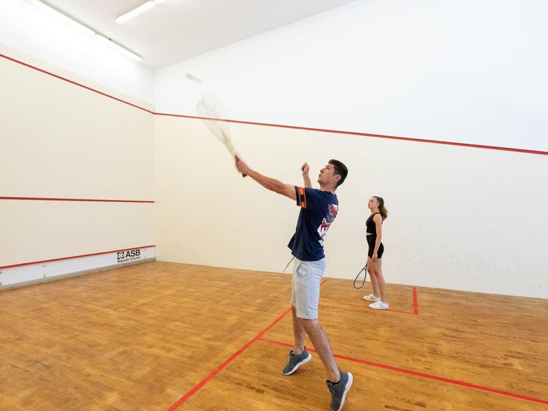 Dva lidé hrají squash na světlém kurtu s bílými stěnami a červenými čarami.