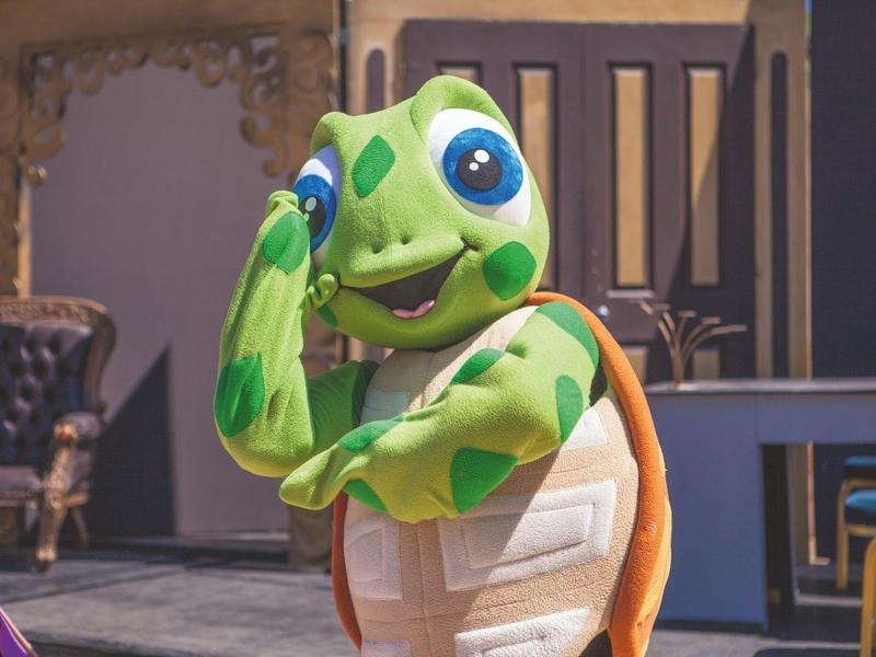 Živá zelená postava želvy před hotelovou terasou s barevným nábytkem.