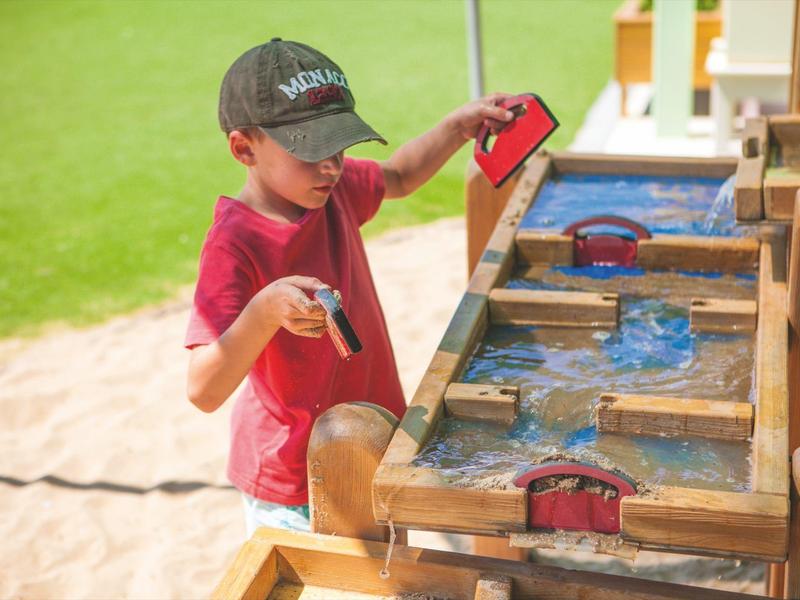 Dítě si hraje na venkovním dřevěném vodním herním prostoru s pískem v pozadí.