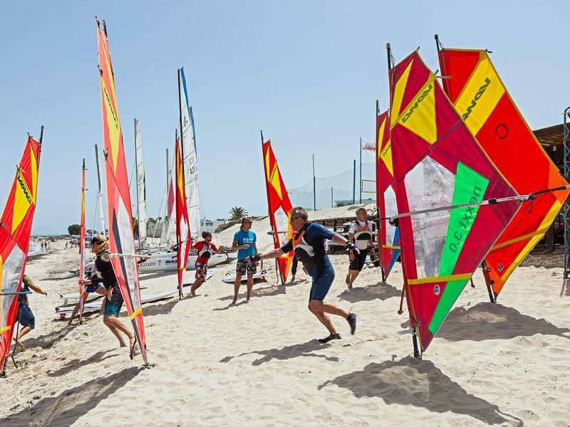 Lidé na pláži připravují barevné windsurfingové prkna.