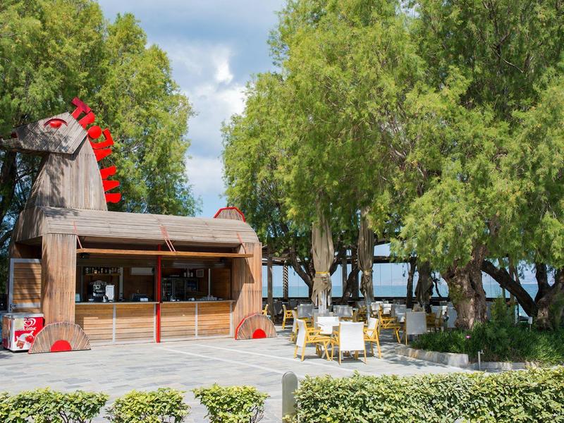 Dřevěná terasa se židlemi pod zelenými stromy u slunečné pláže.