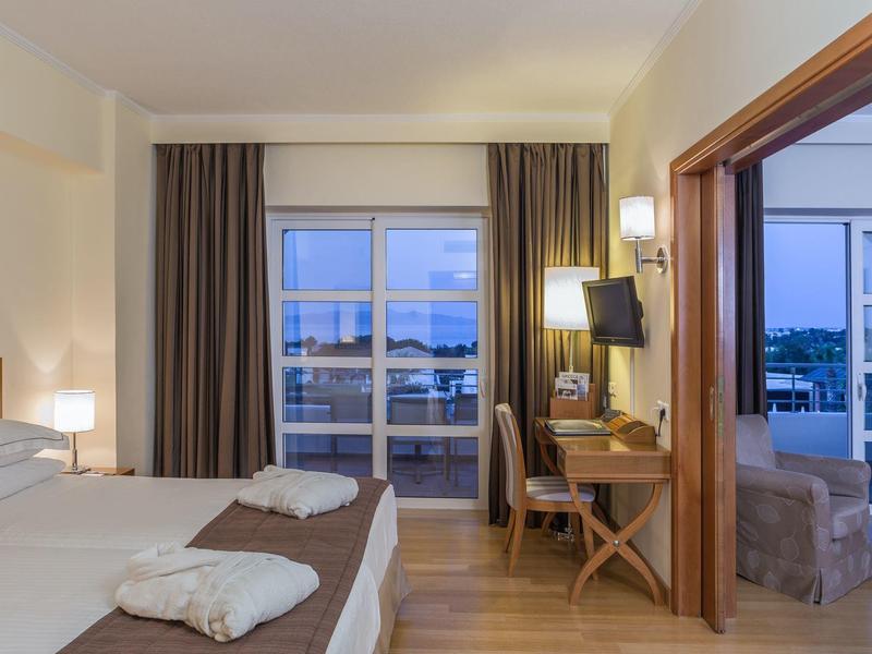 Hotelový pokoj s manželskou postelí, psacím stolem, terasou a výhledem na vodu za soumraku.