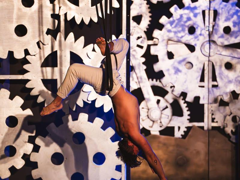 Umělec visící ve vystoupení pole dance před velkými bílými ozubenými koly.