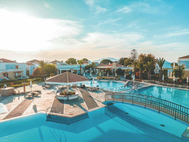 Velký hotelový bazén s slunečníky, lehátky a okolními budovami při západu slunce.