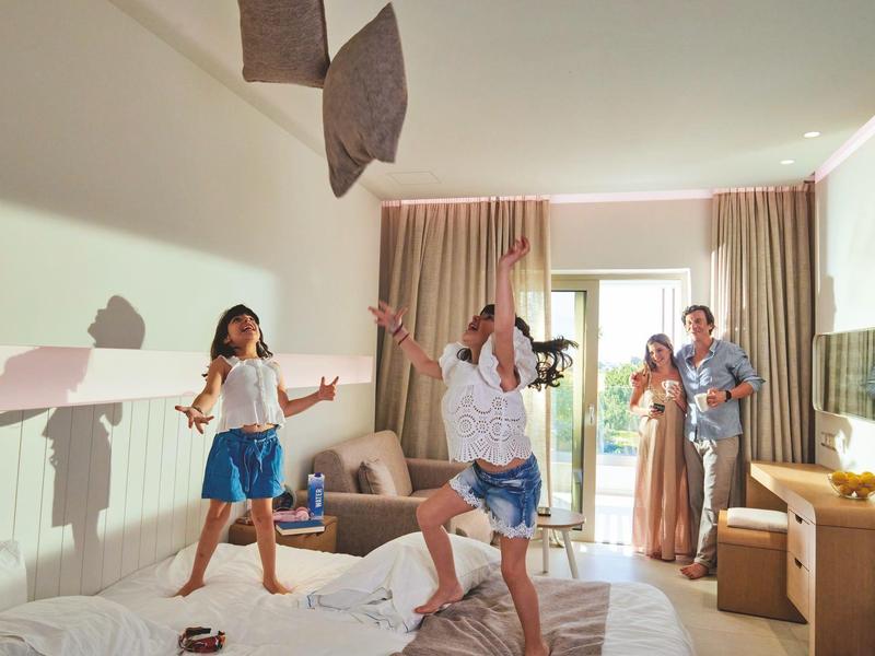 Děti a rodiče radostně skáčou na posteli v hotelovém pokoji.