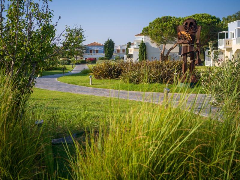 Zpevněná cesta v zelené zahradě obklopená bílými budovami pod modrou oblohou.