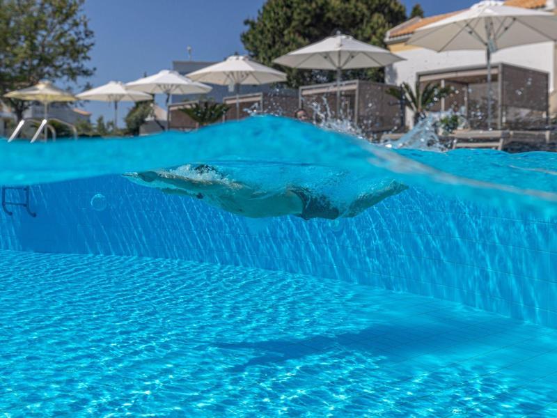 Průzračný bazén s plavcem, bílé slunečníky a budovy v pozadí.