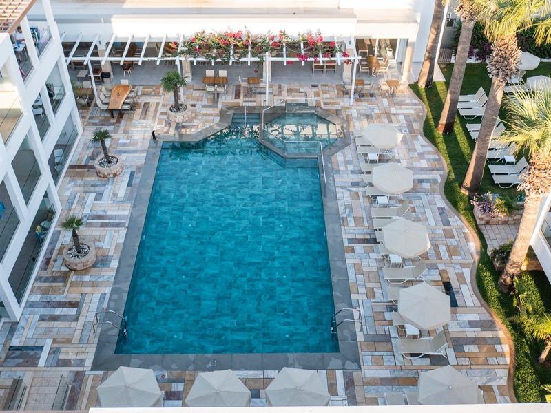 Blick von oben auf einen rechteckigen Pool mit Liegestühlen und weißen Gebäuden daneben.