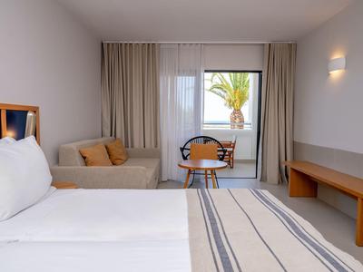 Modernes Hotelzimmer mit großem Bett, Sofa, Tisch und Stühlen, Blick auf Palme durch Fenster.