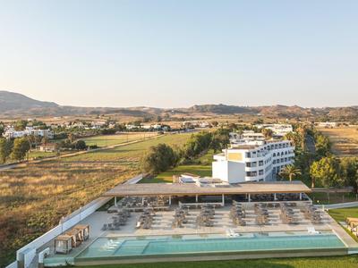 Weißes Hotelgebäude mit Pool und Liegestühlen, umgeben von Feldern und Hügeln bei Sonnenlicht.