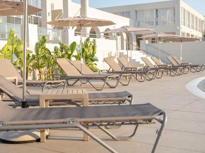 Leere Sonnenliegen am Pool eines Hotels an einem sonnigen Tag mit weißen Gebäuden im Hintergrund.