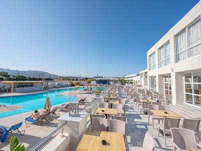 Große Terrasse mit Tischen und Stühlen neben einem Pool und modernem Hotelgebäude bei klarem Himmel.