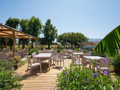 Freiluftrestaurant mit Tischen und Stühlen auf Holzterrasse, umgeben von Grün und blauem Himmel.