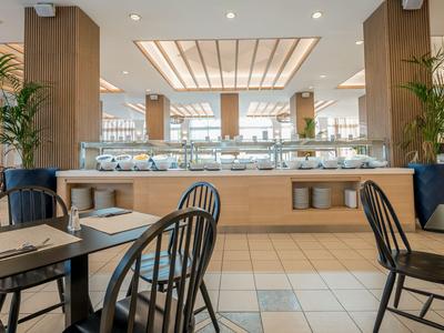Moderner Hotelrestaurant-Speisesaal mit Holztischen, Stühlen und Buffetbereich im Hintergrund.