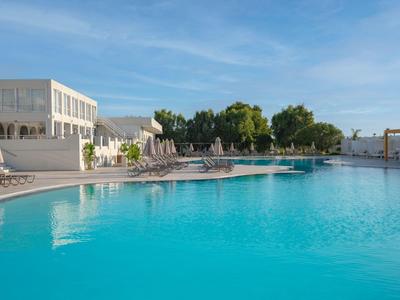 Großer Hotelpool mit Liegestühlen und weißem Gebäude unter blauem Himmel.