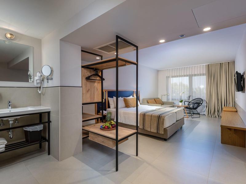 Bright hotel room with modern bed, bathroom area, and seating by the window.