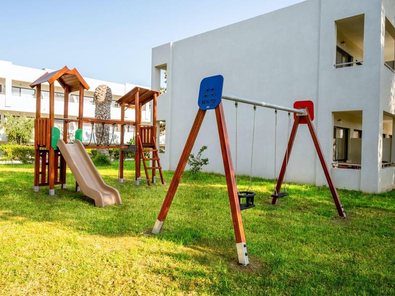 Children's playground with swing and slide next to a white hotel building on a green lawn.