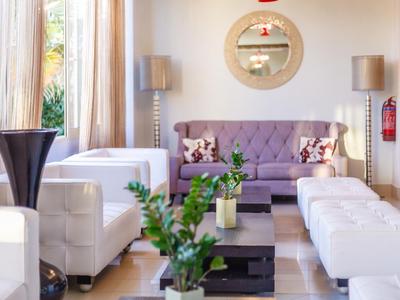 Sala luminosa con sillones blancos, sofá morado y plantas verdes frente a grandes ventanas.