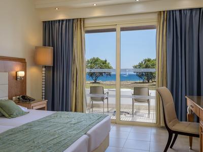 Habitación de hotel con cama doble, escritorio, silla y balcón con vista al mar y cortinas azules.