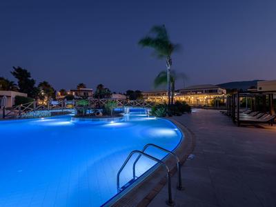 Piscina iluminada en un complejo hotelero durante la noche con palmeras y áreas de descanso.
