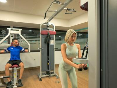 Innenraum eines Fitnessstudios mit zwei Personen, die an Geräten trainieren, helle Wände, Holzboden.