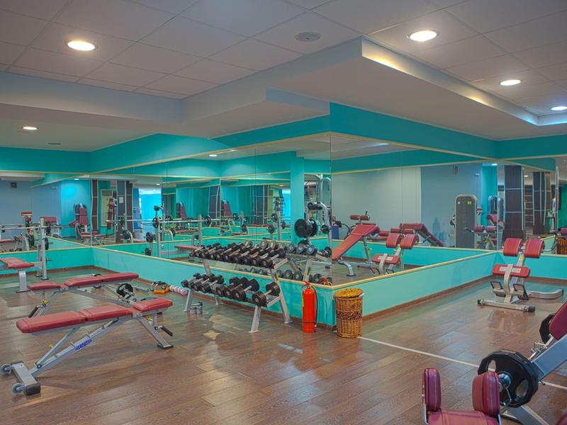 Gimnasio en un hotel con pesas, equipos de entrenamiento y grandes espejos en las paredes.