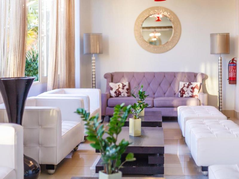 Sala luminosa con sillones blancos, sofá morado y plantas verdes frente a grandes ventanas.