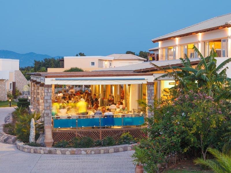 Hotel moderno con terraza iluminada junto a piscina, rodeado de vegetación y montañas al fondo.