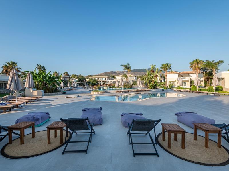 Gran piscina al aire libre con tumbonas y mesas pequeñas, rodeada de villas mediterráneas y cielo azul.