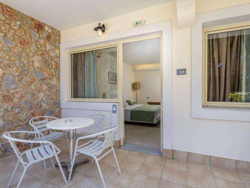 Terraza con mesa de jardín blanca y tres sillas junto a una habitación con puerta y ventana abiertas en un hotel.