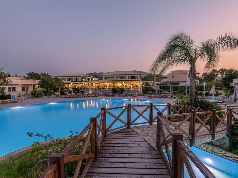 Vista de una piscina de hotel con puente de madera y palmeras al atardecer.