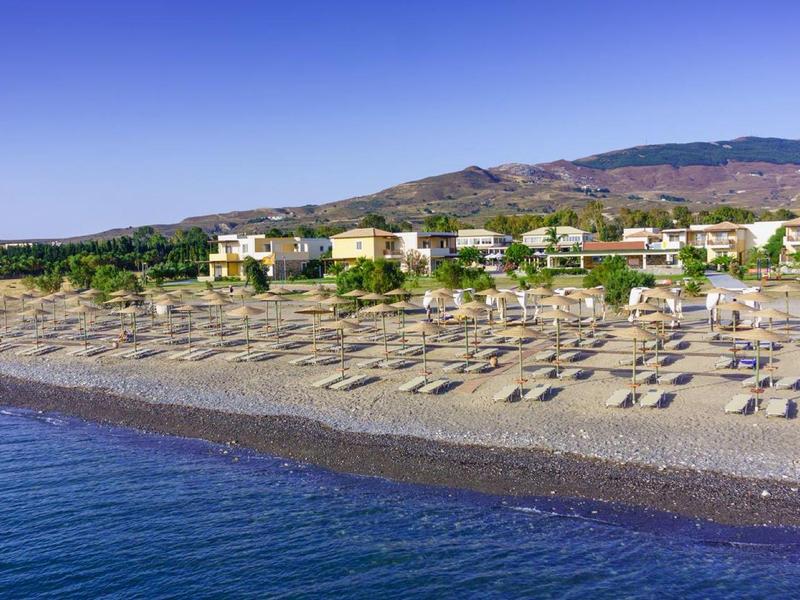 Playa con sombrillas y tumbonas frente a un hotel bajo colinas arboladas junto al mar.