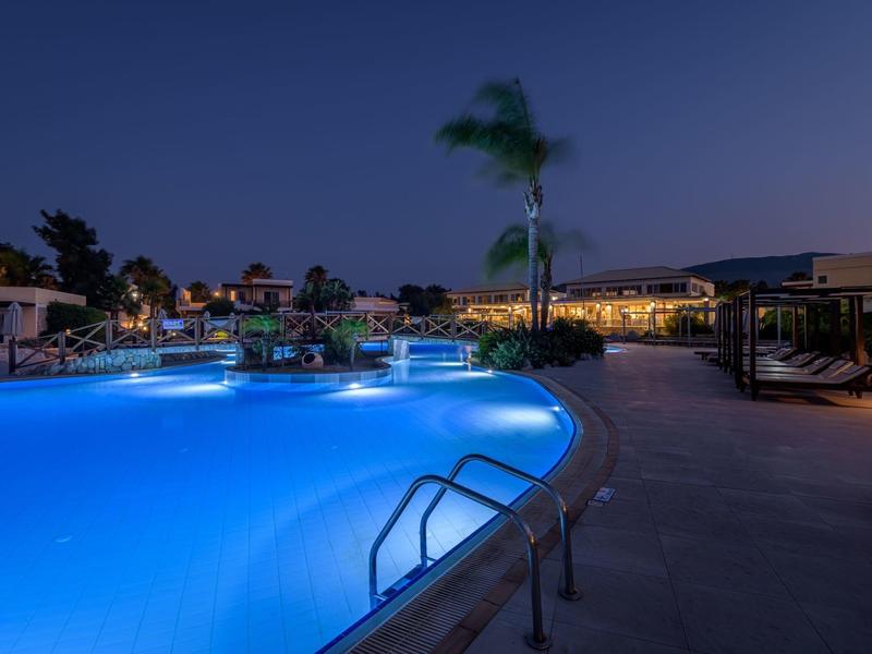 Piscina iluminada en un complejo hotelero durante la noche con palmeras y áreas de descanso.