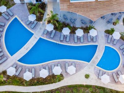 Vista aerea di una piscina curva con lettini e zone verdi in un hotel.