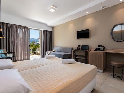 Jasny pokój hotelowy z dużym łóżkiem, sofą, telewizorem i balkonem z widokiem na góry.