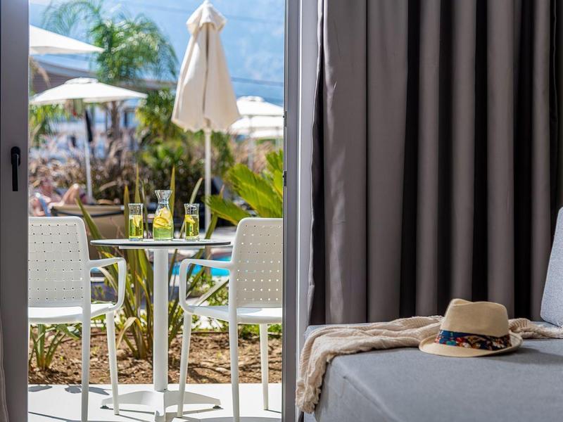 Otwarta szklana drzwi pokazuje taras z krzesłami, stołem i parasolami; wnętrze z sofą i kapeluszem.